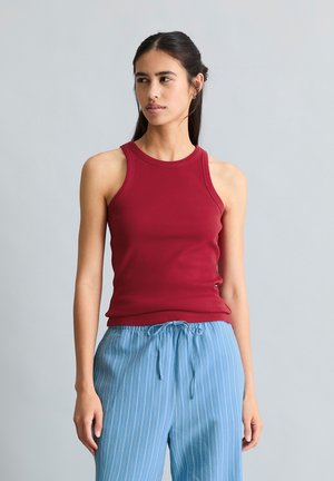 Kobieta o długich ciemnych włosach, ubrana w czerwoną bluzkę bez rękawów i jasnoniebieskie, paskowane spodnie ze sznurkiem, patrzy w prawo.