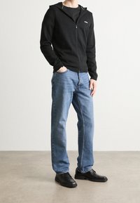 Schwarze Zip-Hoodie mit geripptem Design und grünen Akzenten, getragen über einem schwarzen T-Shirt, kombiniert mit hellblauen Jeans und schwarzen Schuhen.