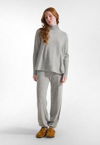 Grau, gerippter Rollkragenpullover kombiniert mit einer passenden, lockeren Hose. Braune, fusselige Hausschuhe runden das Outfit ab. Weicher, strukturierter Stoff.