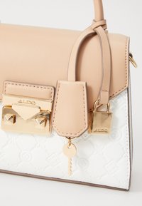 Sac à main en cuir beige avec accessoires dorés, présentant un fermoir triangulaire, un charme en forme de clé, et une base blanche texturée avec un motif en relief.