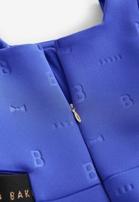 Mėlyna audinio suknelė su reljefiniais "B" ir kaspino raštais. Turtinga auksine užtrauktuko traukle ir tekstūruotu paviršiumi. Išskirtinis dizainas.
