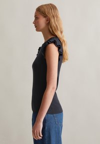 Zwarte geribde top met ruches aan de mouwtjes, slim fit en een gladde textuur. Model draagt het met een blauwe denim onderkant, waarbij een zijaanzicht wordt getoond.