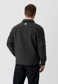 Giacca di lana grigio scuro con colletto, dotata di un'etichetta quadrata sul retro e maniche textured, abbinata a pantaloni in denim scuro.