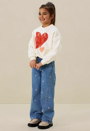 Jeune fille souriante, portant un sweat-shirt blanc avec des cœurs rouges, un jean bleu avec de petits imprimés de fleurs blanches et des chaussures beiges avec des semelles noires.
