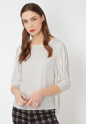 Mujer con cabello largo castaño que lleva una blusa blanca de mangas de flecos y pantalones a cuadros en blanco y negro, de pie contra un fondo blanco liso.