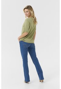 Top scurt cu mânecă scurtă de culoare verde măslin, cu puncte texturate, asortat cu blugi evazați albaștri și tocuri bej, văzut din spate. Design simplu.