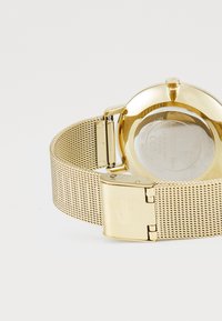 Montre avec bracelet en maille dorée, dotée d'un boîtier rond, d'une finition polie et d'une fermeture à pression. L'arrière présente des détails de marque et une surface lisse.