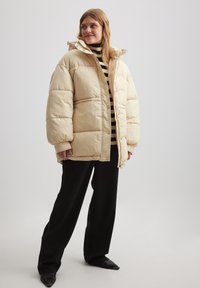 Žena v béžovej prešívanej bunde, čiernom a béžovom pruhovanom roláku, čiernych širokých nohaviciach a čiernych špicatých topánkach, stojaca na neutrálnom pozadí.