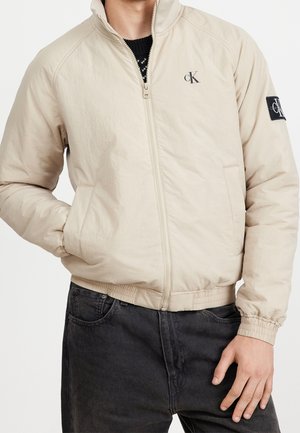 Man draagt een beige jas met ritssluiting met CK-logo op borst en mouw, gecombineerd met een zwarte jeans, hand in zak en ontspannen houding.