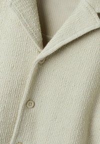 Strukturerad, off-white jacka med en spetsig krage, knappstängning och synliga sömmar för extra detaljer.