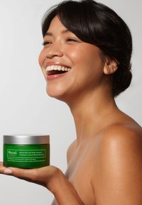 Murad Retinal ReSculpt Body Treatment i en grön glasburk med ett silverlock. Burkens etikett har vit text som beskriver produkten. Modell håller i burken.