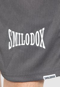 Pantalones cortos de canalé grises con "SMILODOX" en texto blanco en negrita en la pierna, con una etiqueta de tela que también muestra el nombre de la marca.