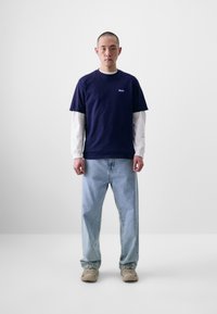 Un tricou din bumbac bleumarin suprapus peste o cămașă albă cu mânecă lungă, asortat cu blugi deschiși la culoare și adidași bej, pe un fundal neutru.