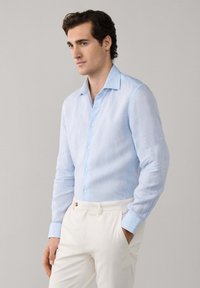 Junger Mann, der ein hellblaues, langärmliges Hemd mit Knöpfen und weiße Hosen trägt, mit einer Hand in der Tasche vor einem einfarbigen Hintergrund steht.