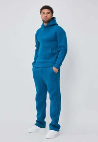 Sweat à capuche bleu avec poche kangourou et pantalon assorti, fabriqués dans un tissu doux. Comprend des poignets côtelés et une coupe décontractée. Baskets blanches.