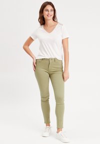 Haut blanc à manches courtes et en v associé à un jean vert olive ajusté. Le mannequin porte des baskets blanches. Design simple, texture douce et coupe slim.