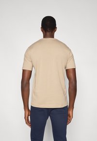 Μπεζ βαμβακερό t-shirt με κοντά μανίκια και στρογγυλή λαιμόκοψη, εμφανιζόμενο από πίσω, σε συνδυασμό με σκούρο μπλε παντελόνι. Ομαλή υφή, κανονική εφαρμογή.