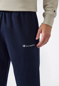 Mano derecha descansando cerca de unos pantalones de chándal azul marino con el logo blanco de Champion, sudadera de manga larga beige con puños que muestra un pequeño logo en la manga.