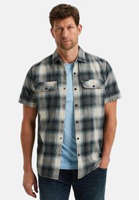 Korte mouwen geruite shirt in zwart, wit en blauw; heeft twee borstzakken, knoopsluiting en een comfortabele pasvorm, gedragen over een lichtblauw T-shirt.