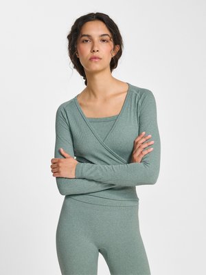 Kvinde med fletninger iført en ribstrikket, salviegrøn omslagsbluse med lange ærmer og matchende tætsiddende bukser, stående med armene krydset.