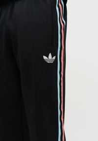 Pantalon de sport noir avec des bandes latérales roses et bleu clair, et un logo trèfle Adidas blanc près de la poche.