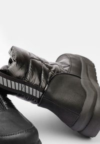 Bottines noires avec un dessus matelassé et brillant et une base en cuir texturé. Équipées d'un détail élastique rayé le long de la tige et d'une semelle épaisse.