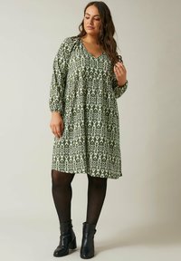 Grünes gemustertes Kleid mit V-Ausschnitt und langen Ärmeln, das ein fließendes Design und eine taillierte Taille aufweist, kombiniert mit schwarzen Stiefeletten.