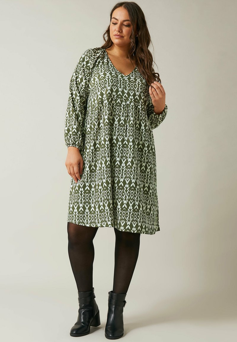 Grünes gemustertes Kleid mit V-Ausschnitt und langen Ärmeln, das ein fließendes Design und eine taillierte Taille aufweist, kombiniert mit schwarzen Stiefeletten.