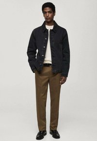 Dunkelblaue Jeansjacke mit Standardkragen, Knopfverschluss und langen Ärmeln, kombiniert mit einem hellen Pullover und braunen Hosen; getragen mit schwarzen Schuhen.