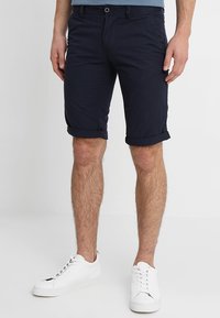 Pantaloni corti blu navy in cotone con orlo arrotolato, dotati di tasche laterali, chiusura frontale con bottone e scarpe da ginnastica bianche abbinate.