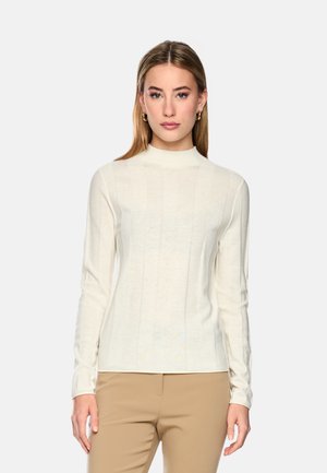 Young woman with straight light brown hair wears a fitted long-sleeve ivory top and beige pants against a plain light background.
