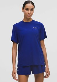 Fiatal nő kék, rövid ujjú pólót és passzoló sortot visel, egy egyszínű világos háttér előtt áll.