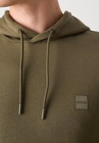 Gros plan d'une personne portant un sweat à capuche vert olive avec cordons et un petit patch rectangulaire avec le logo "BOSS" sur la poitrine.