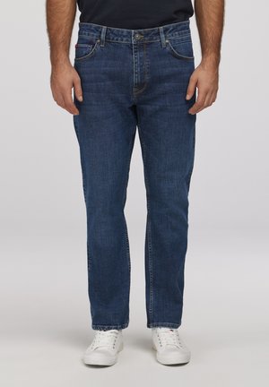 Mężczyzna noszący ciemnoniebieskie jeansy o prostych nogawkach i białe tenisówki, stojący na tle jednolitego jasnoszarego tła.