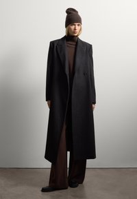 Vrouw in een bruine gebreide hoed, donkerbruin coltrui, wijde broek, lange zwarte overjas en zwarte schoenen, staande binnenshuis.