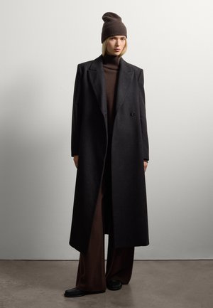 Vrouw in een bruine gebreide hoed, donkerbruin coltrui, wijde broek, lange zwarte overjas en zwarte schoenen, staande binnenshuis.