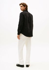 Zwarte langärmelige shirt met knoopmanchetten, voorzien van een achterkant met schoudernaden en een rechte snit, gecombineerd met witte rechte broek en zwarte schoenen.