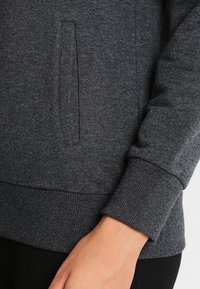 Sudadera gris oscuro con una textura suave, bolsillo tipo canguro, puños acanalados y diseño ajustado en el dobladillo.