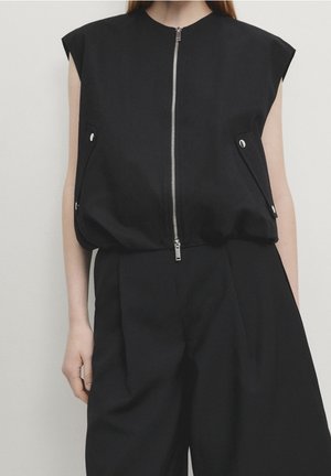 Personne portant un haut noir sans manches zippé avec des poches à rabat avant et un pantalon noir à jambes larges assorti sur un fond uni.
