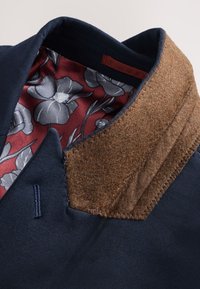 Marineblauem Stoff mit einem braunen Filzkragen. Das Innere zeigt ein rotes Blumenmuster auf grauem Hintergrund. Sichtbare Nähte entlang der Kanten.