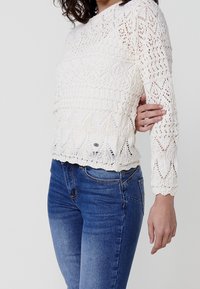 Bílý háčkovaný svetr s texturovaným vzorem obsahujícím diamantu a obloučkové motivy. Dlouhé rukávy a přiléhavý střih. Kombinován s modrými džínami.