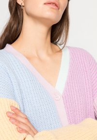 Pastel-colored cardigan with a v-neck, featuring a blue, purple, and yellow striped pattern and a pink button detail. Knitted texture visible.
