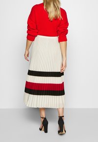 Jersey de punto rojo, falda midi plisada en crema con rayas horizontales negras y rojas. Zapatos de tacón alto negros completan el conjunto.