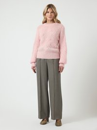 Mulher loira de pé vestindo um suéter rosa de malha canelada, calças largas cinzas e sapatos de salto bejes, contra um fundo branco.