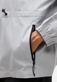 Χέρι στην πλαϊνή τσέπη ενός ανοιχτόχρωμου γκρι μπουφάν με μαύρο φερμουάρ και ελαστικό μανσέτι, με την επιγραφή "DRY-XD" στο μανίκι.