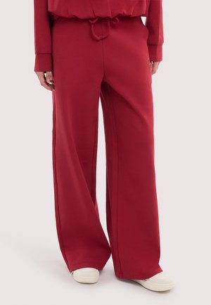 Person iført løstsiddende røde joggingbukser med snøre i taljen og hvide sneakers, stående mod en ensartet lys baggrund.