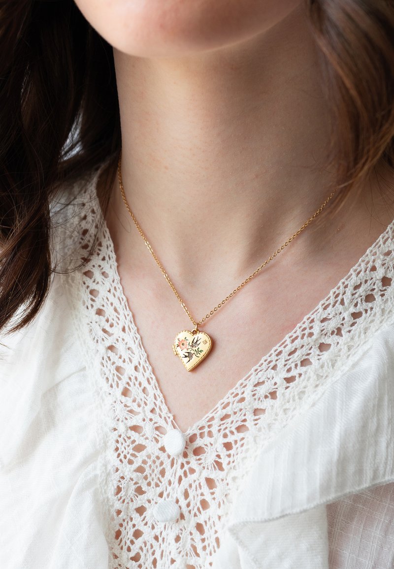 Gouden hartvormige hanger met bloem- en vogelmotief aan een fijne ketting, gedragen over een witte blouse met kanten rand.