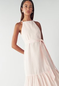 Vestido sin mangas en rayas rosa claro y blanco, ceñido en la cintura con un lazo de tela, con un dobladillo escalonado y una textura suave.