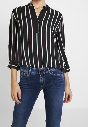 Mujer con una blusa negra con rayas blancas verticales y vaqueros azules, de pie frente a un fondo liso y claro.