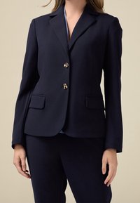 Blazer bleu marine en tissu lisse, doté d'un col à revers cranté, de deux poches avant et d'une double rangée de boutons dorés pour la fermeture.